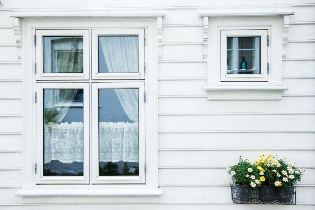 Hvit trehusfasade med klassiske vinduer, blondegardiner og blomsterkasse med gule og hvite blomster